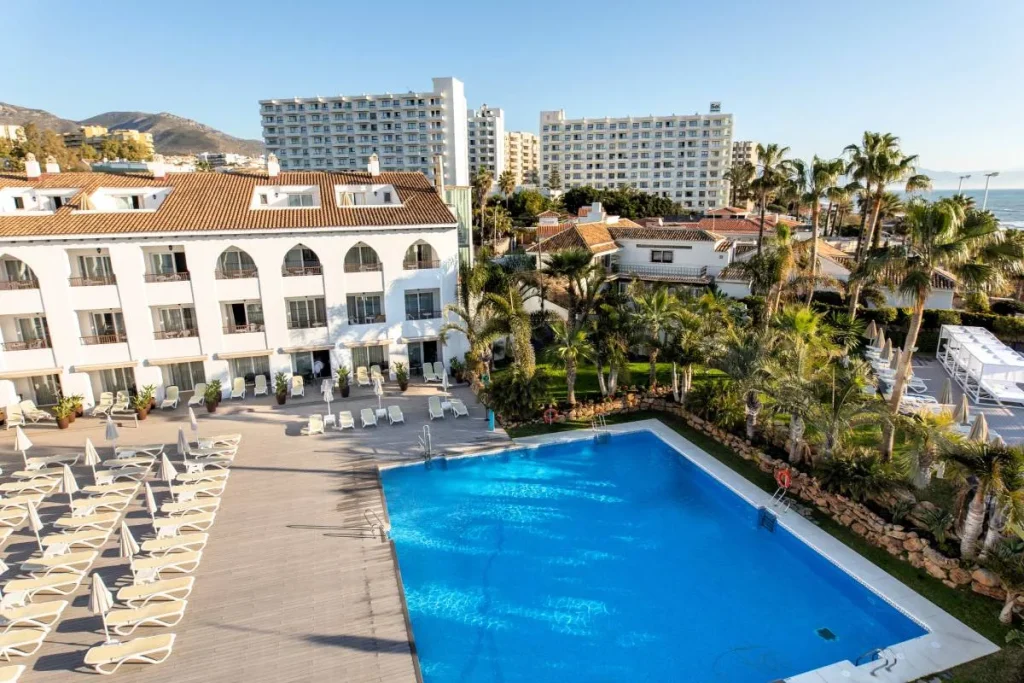 Hotel con piscina azul, tumbonas y palmeras junto al mar y monta&ntilde;as.