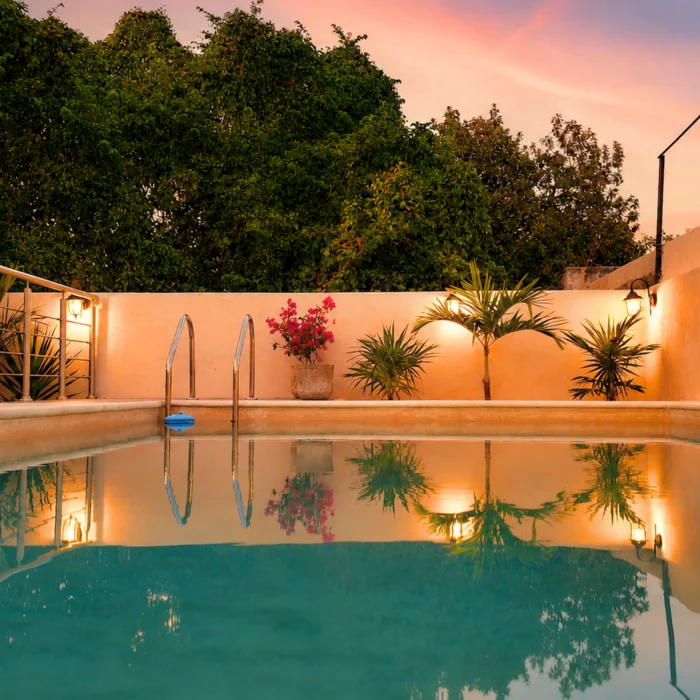 Piscina al atardecer con plantas y flores en un patio con luces c&aacute;lidas.
