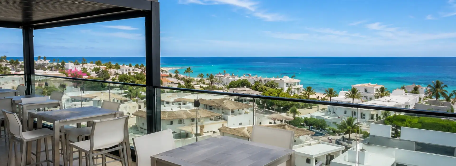 Terraza con mesas y vista al mar turquesa y un pueblo costero con palmeras.