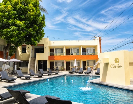 Hotel junto a piscina con tumbonas, &aacute;rboles y cielo azul.