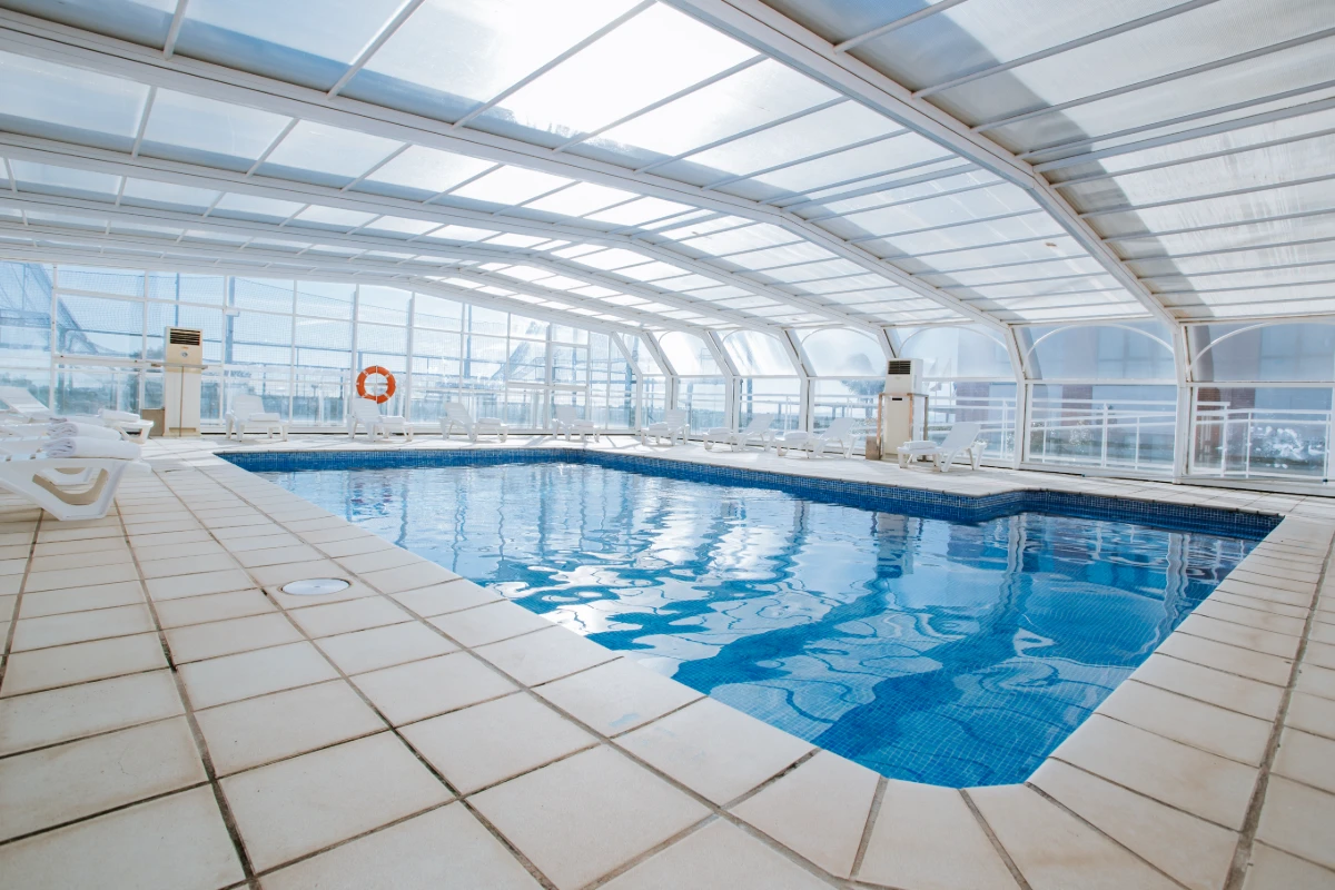 Piscina cubierta con techo de vidrio y tumbonas blancas, vista interior