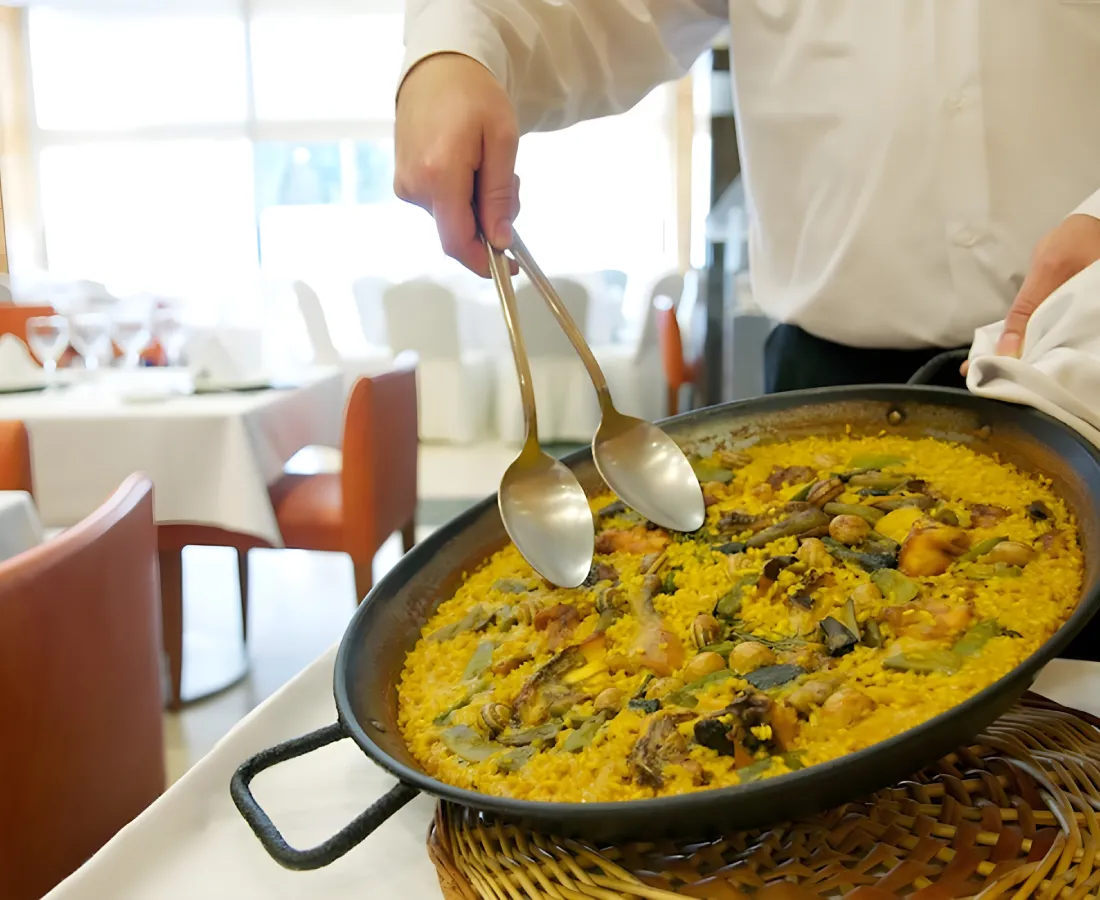 Chef sirviendo paella amarilla en una sart&eacute;n con cucharas doradas.