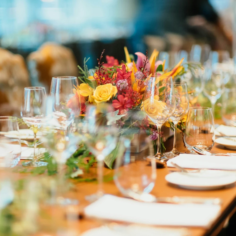 Arreglo floral colorido en mesa de banquete con copas de cristal y ambiente c&aacute;lido
