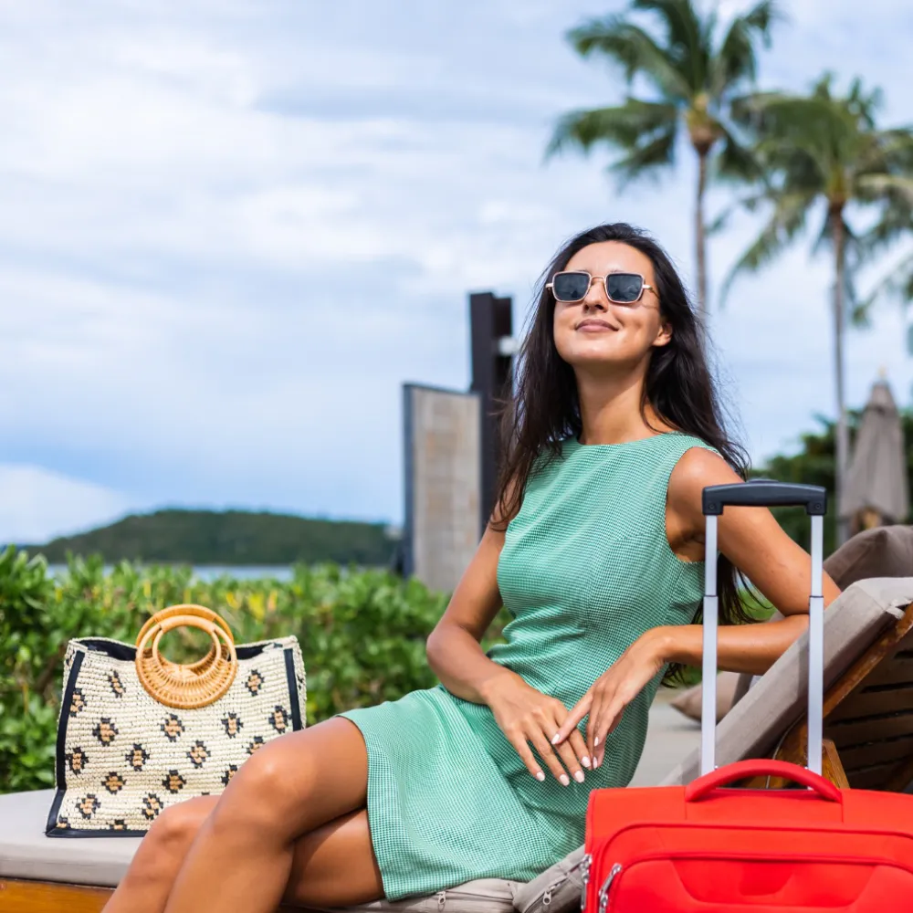 Mujer con gafas de sol en terraza, con bolso de mimbre y maleta roja.