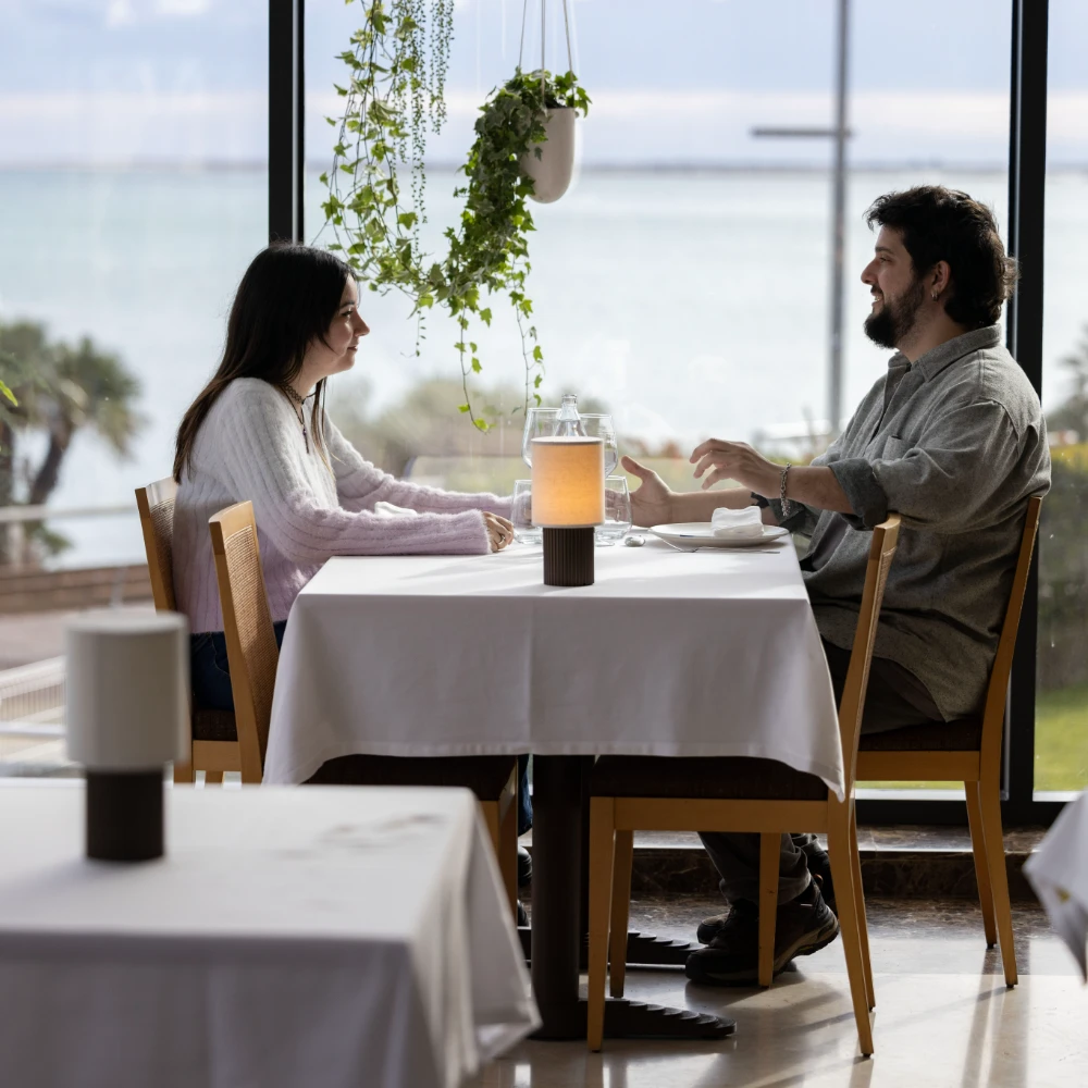 Pareja desayunando en terraza junto a una ventana con plantas y mesa puesta