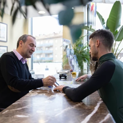 Dos hombres estrechan manos en oficina, con mesa y plantas al fondo.