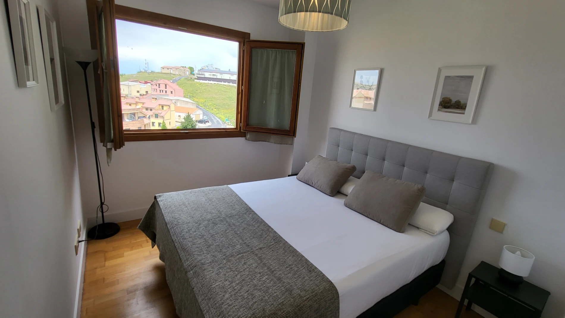 Dormitorio con cama doble, ventanal y cortinas, con vista a un barrio verde