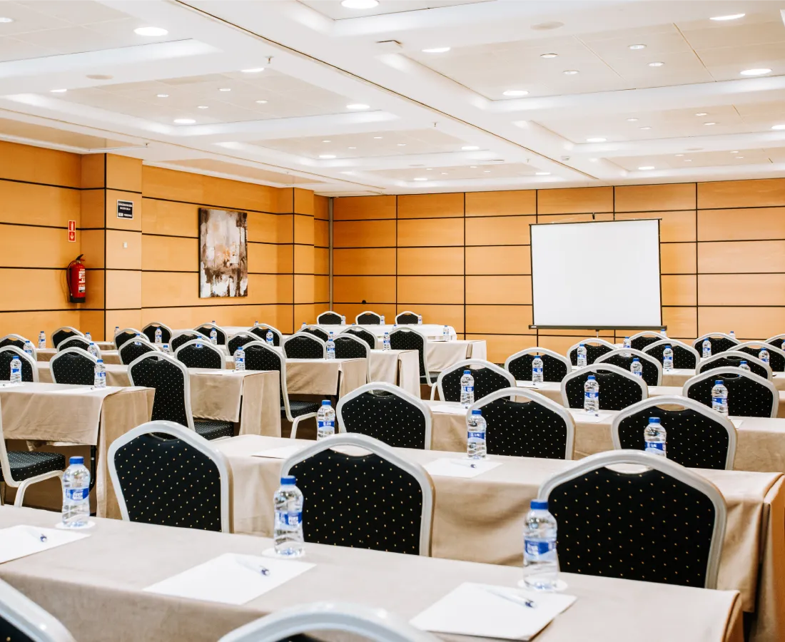 Sala de conferencias con mesas, sillas y botellas de agua al frente.