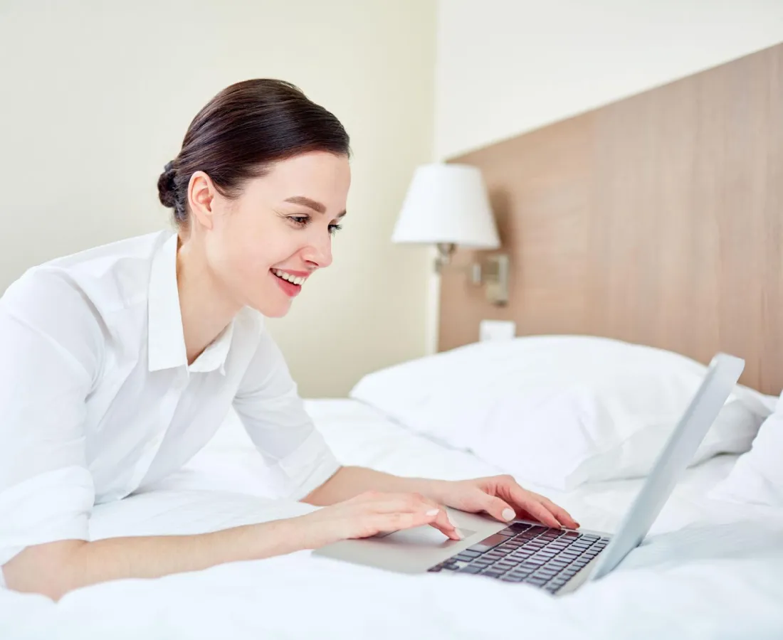 Mujer en cama usando laptop en una habitaci&oacute;n hotelera luminosa