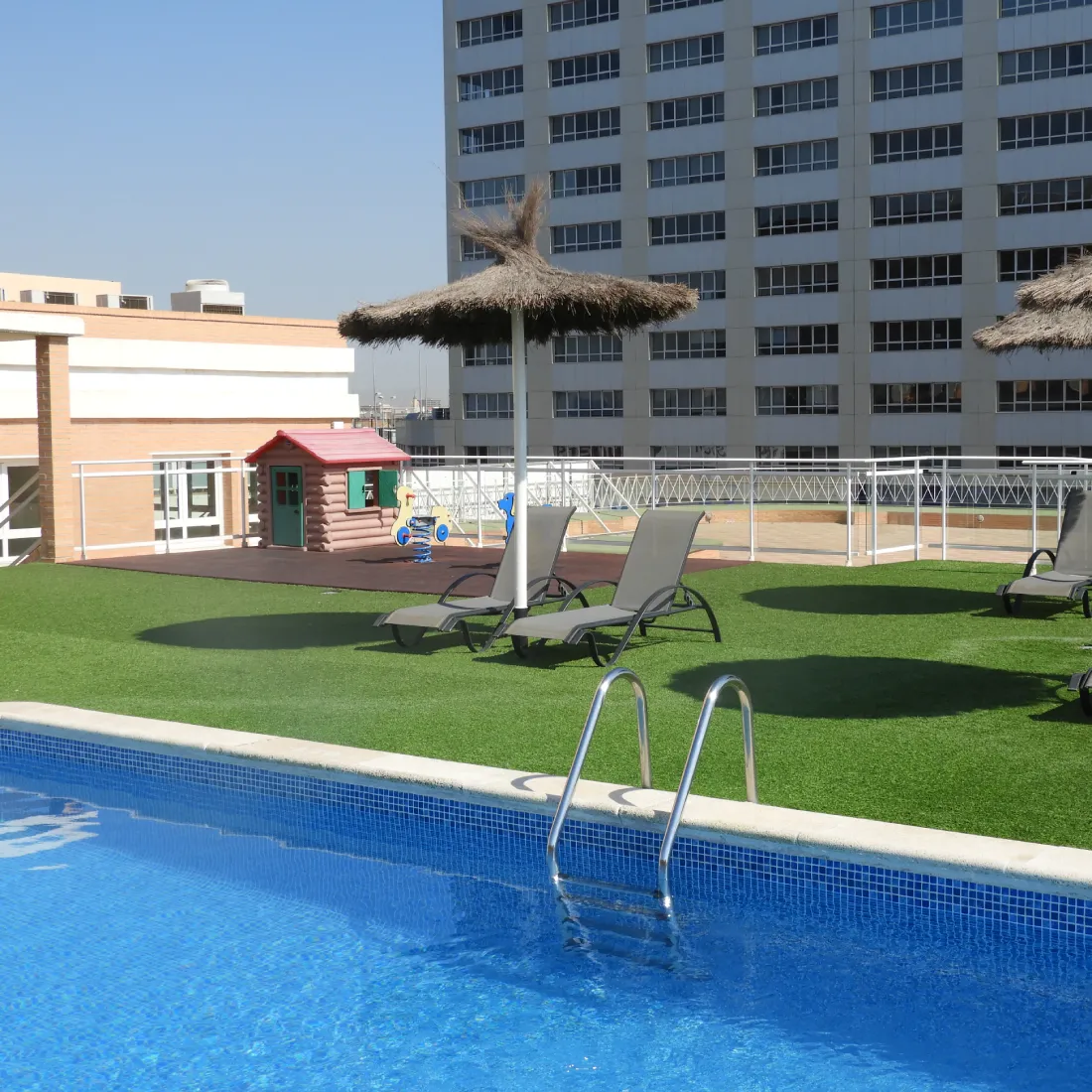 Piscina con c&eacute;sped artificial, tumbonas y casita infantil en patio de hotel