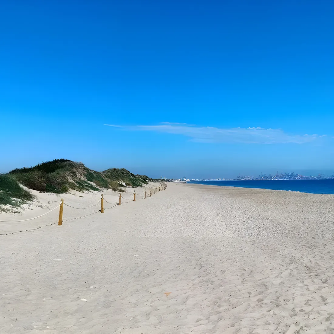 Playa de arena blanca con dunas y vista al puerto en el horizonte