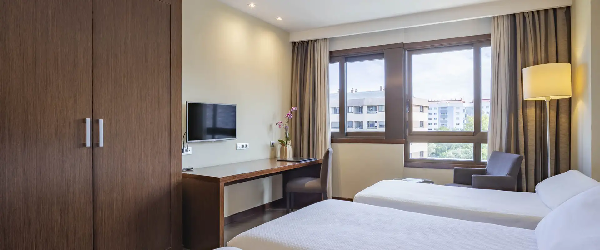 Habitaci&oacute;n hotelera moderna con escritorio, TV, cama y grandes ventanas