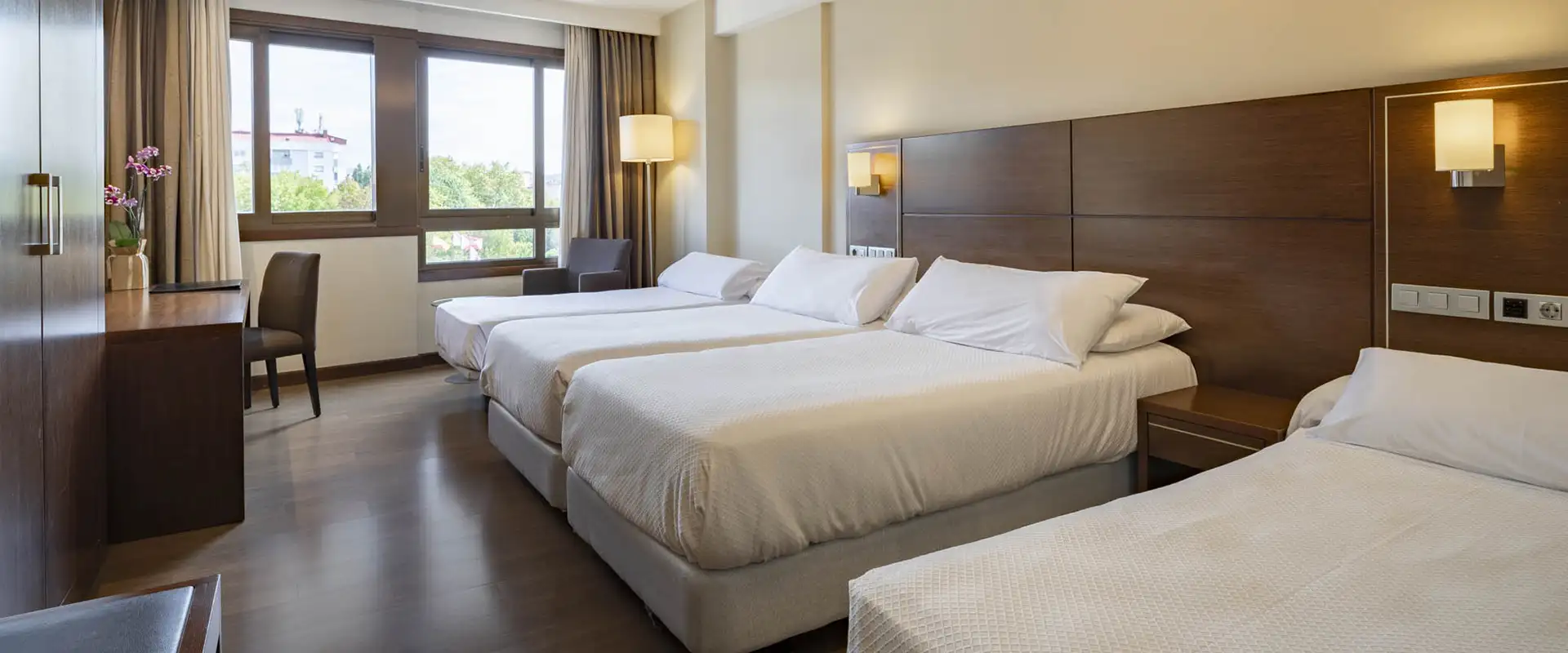 Habitaci&oacute;n de hotel moderna con camas dobles, ventanales y l&aacute;mparas de pared.