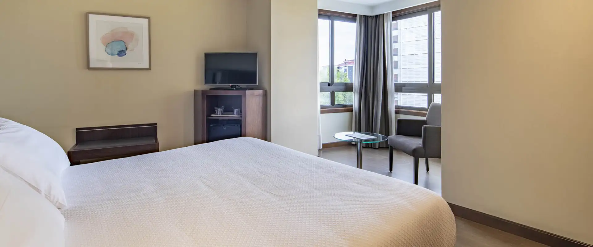 Habitaci&oacute;n de hotel con cama, televisor, mesita y ventana con cortinas.
