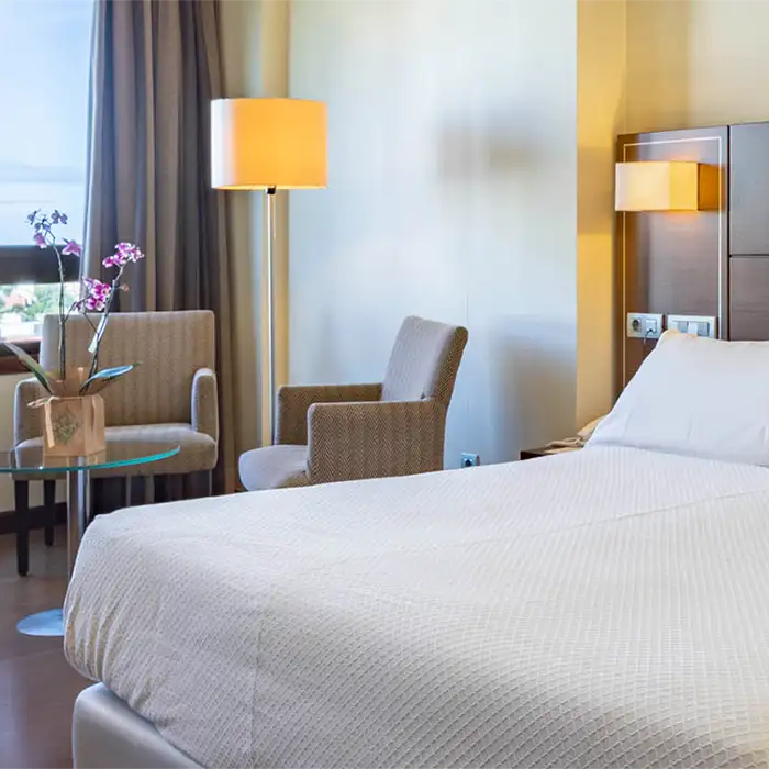 Habitaci&oacute;n de hotel luminosa con cama, sill&oacute;n y l&aacute;mparas de pie.