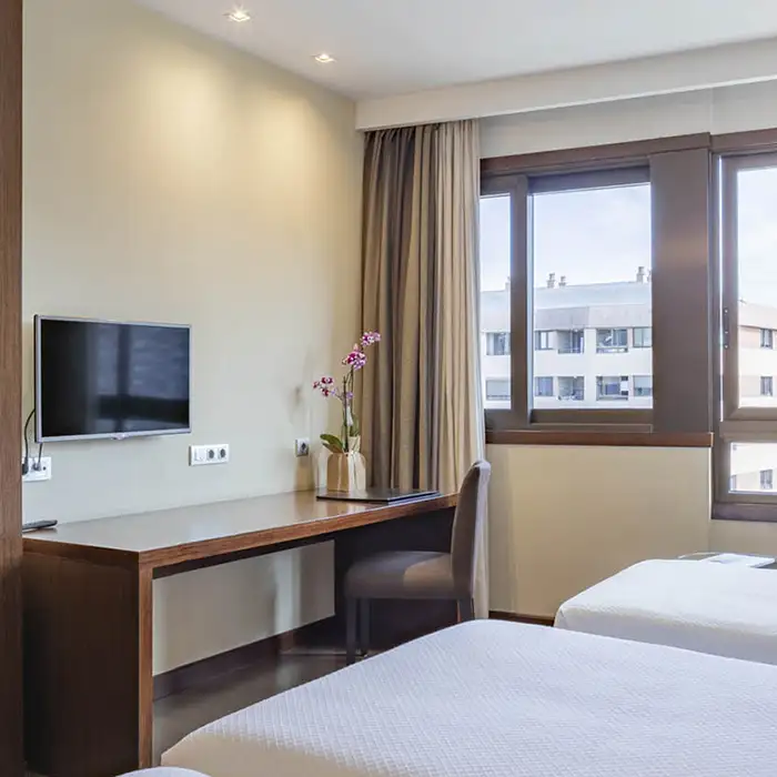 Habitaci&oacute;n de hotel con escritorio, silla, TV y ventana con cortinas.