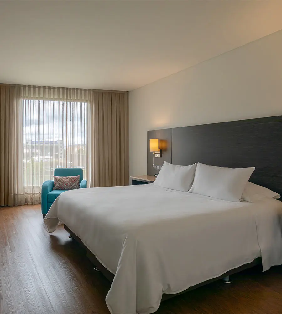 Dormitorio moderno con cama doble, sill&oacute;n azul y cortinas beige junto a la ventana