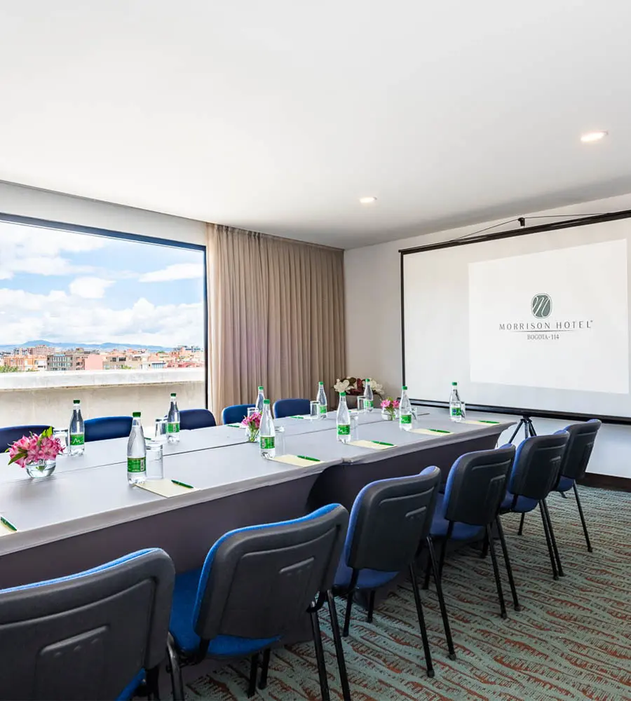 Sala de reuniones en hotel con mesa, sillas azules y pantalla blanca.