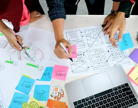 Personas trabajando en una mesa con planos y notas, cerca de una laptop.