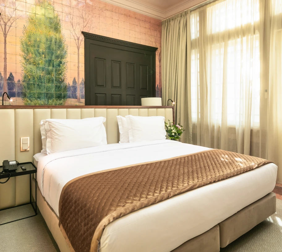 Quarto de hotel com cama e cortinas claras, parede decorada e luz natural