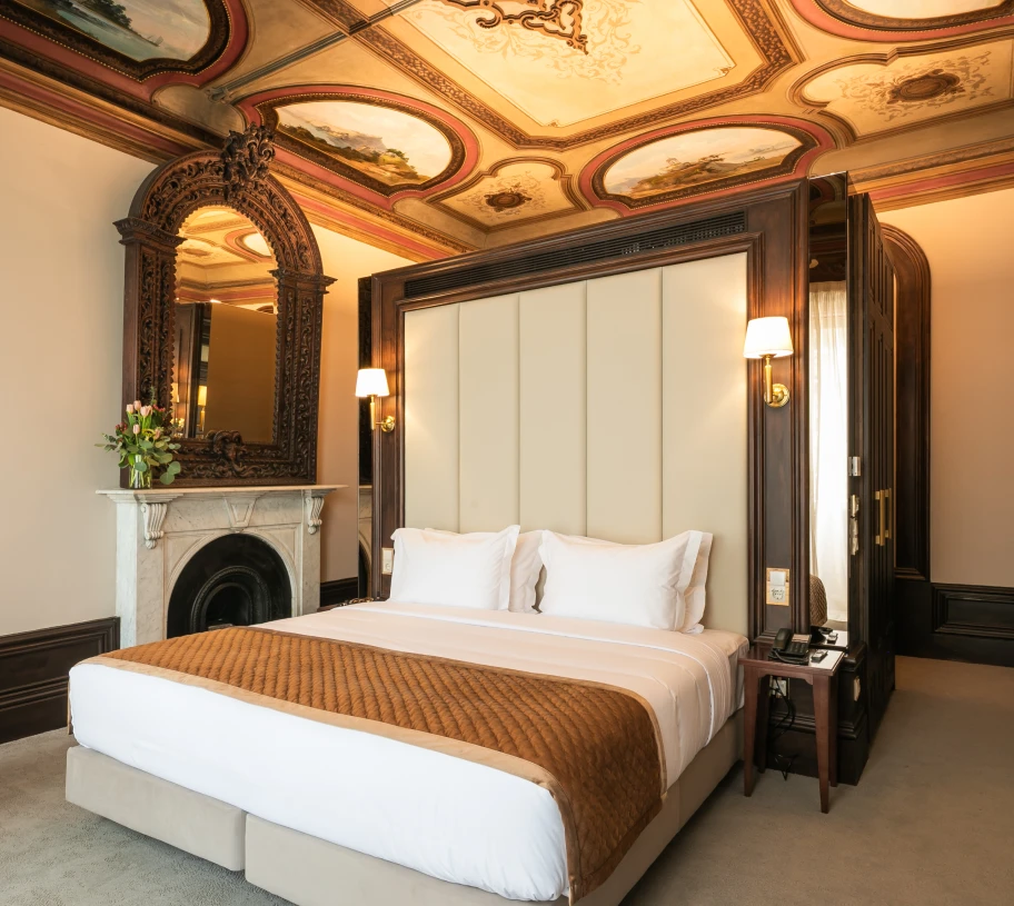 Quarto de hotel cl&aacute;ssico com cama e lareira ao fundo, teto decorado.