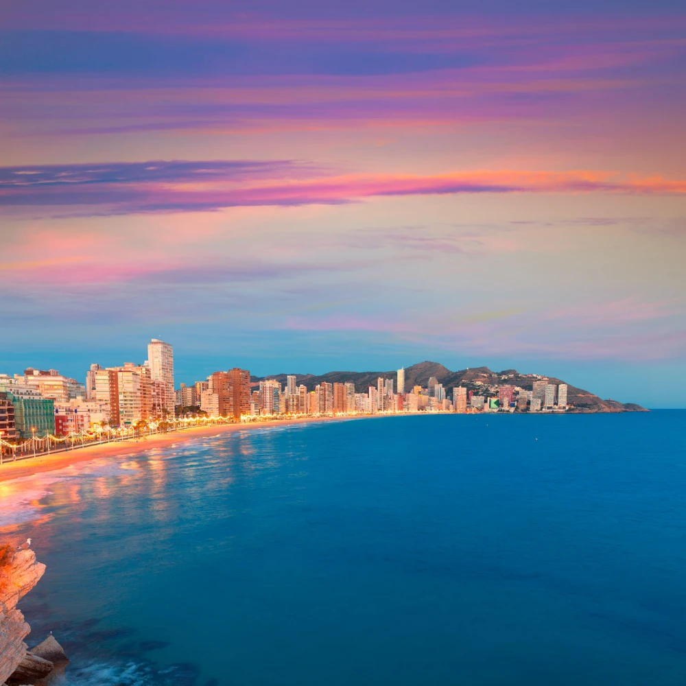 Atardecer sobre un malec&oacute;n costero con edificios y monta&ntilde;as al fondo
