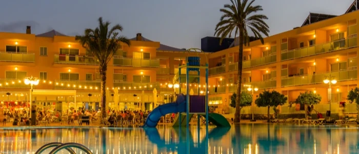 Hotel con piscina y palmeras iluminadas al atardecer, vista desde el agua.