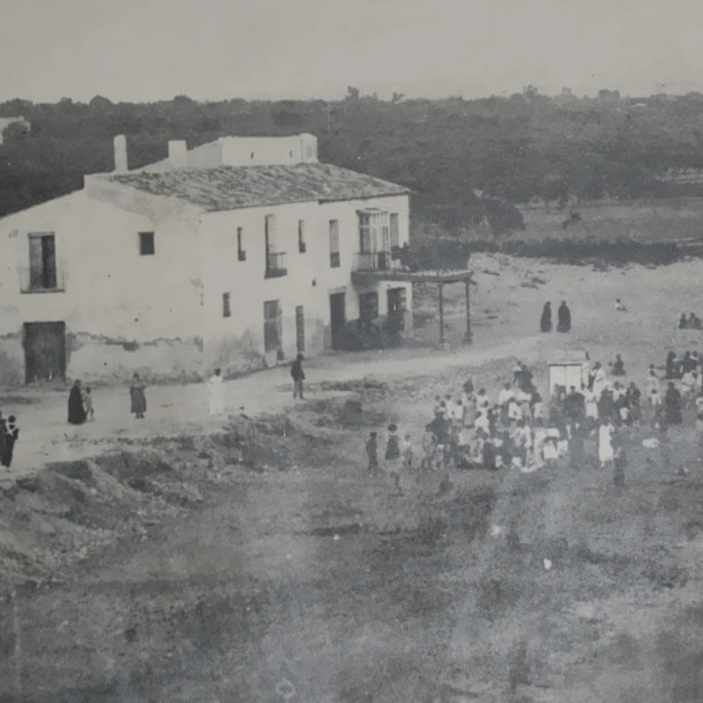 Gente reunida frente a un edificio blanco junto a un camino rural, al fondo un bosque
