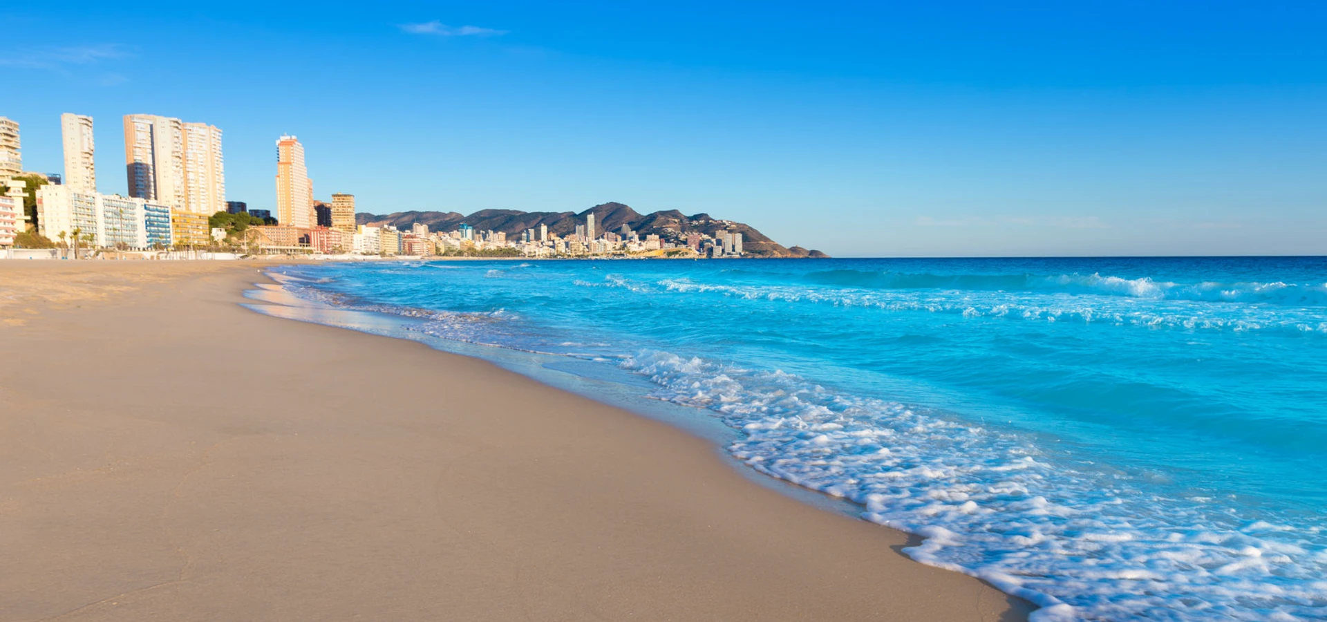 Playa de arena con olas azules y edificios al fondo bajo cielo despejado