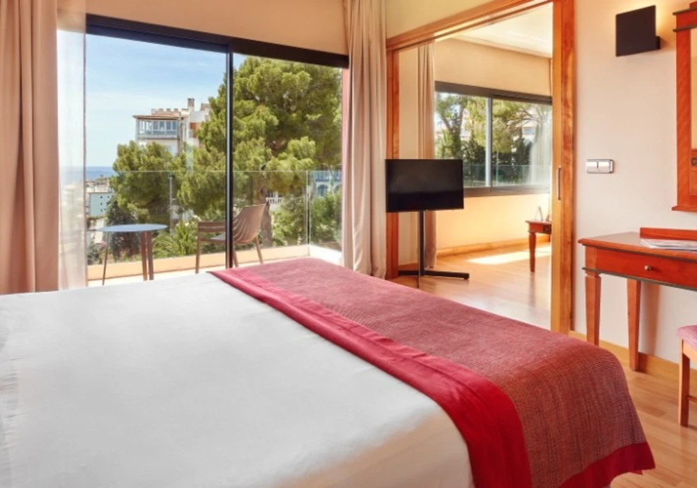 Dormitorio con cama de s&aacute;banas blancas y manta roja, con vista al mar