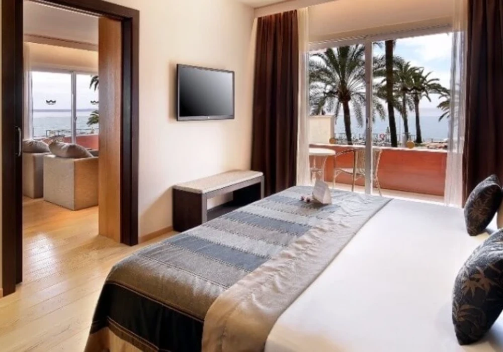Dormitorio moderno con cama, televisi&oacute;n y ventanales con vista al mar y palmeras