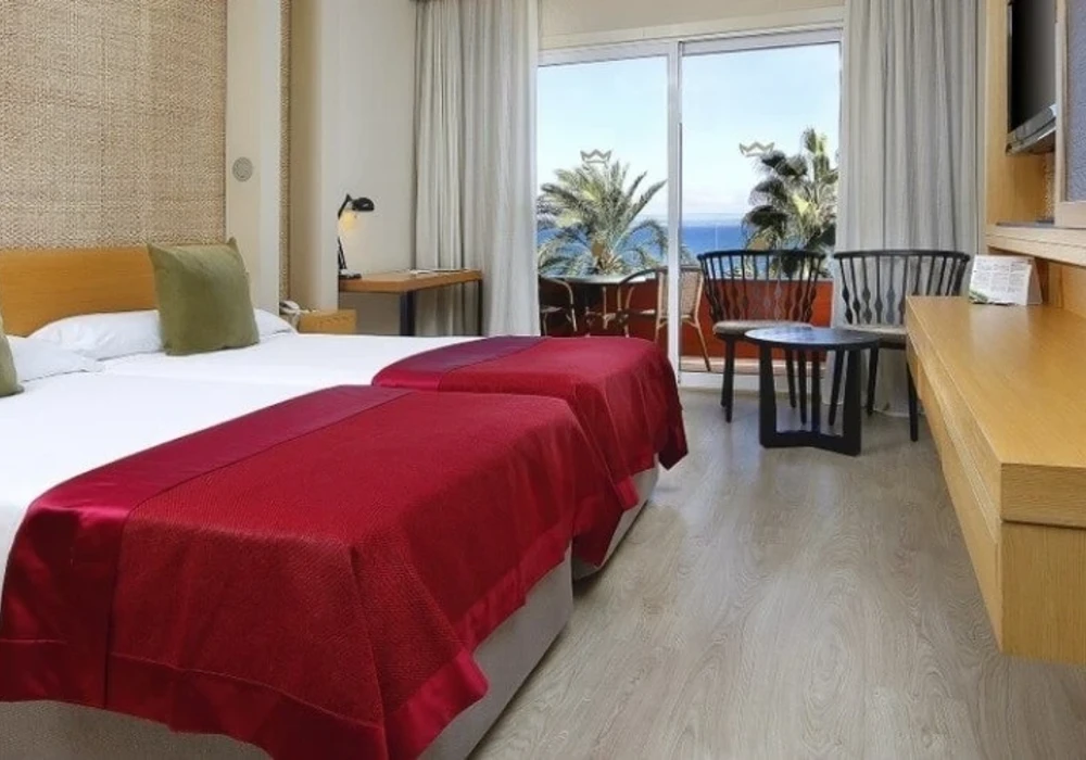 Habitaci&oacute;n de hotel con cama roja, mesas y vista al mar por la ventana