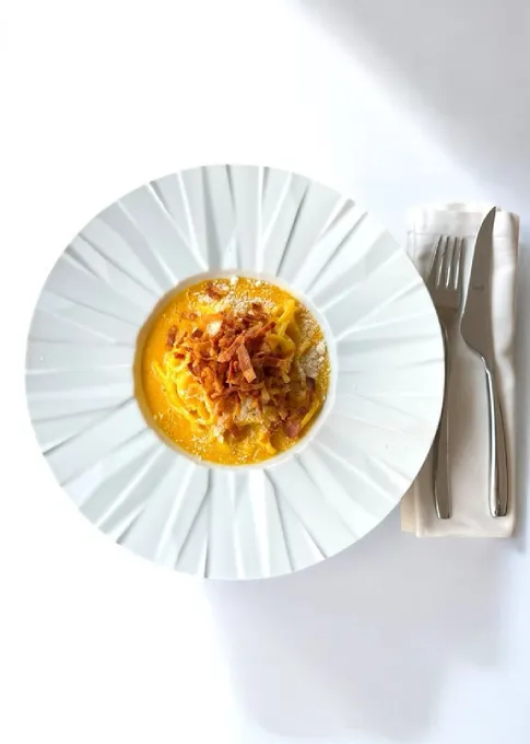 Plato con crema amarilla y tocino sobre fondo blanco, con cubiertos.