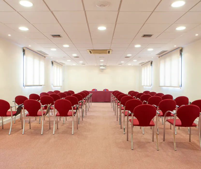 Sala de conferencias con filas de sillas rojas y mesa central vac&iacute;a