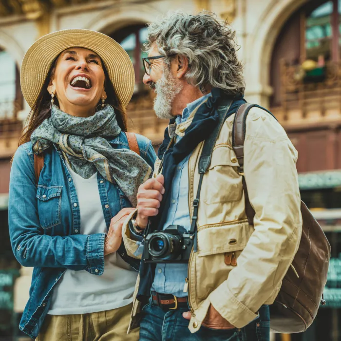 Pareja de turistas con sombrero y c&aacute;maras, riendo frente a una calle.