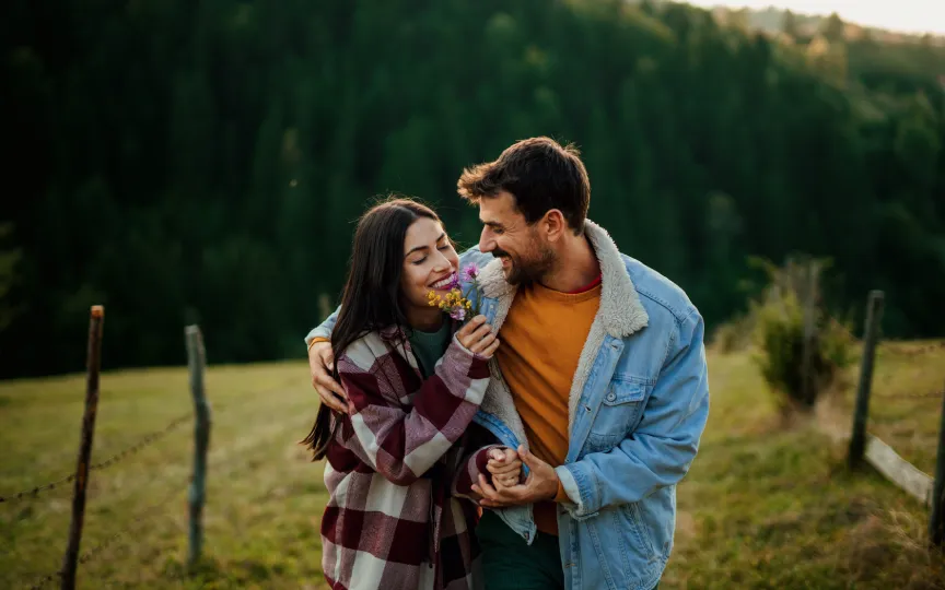 Pareja en el campo abraz&aacute;ndose mientras ella sostiene flores.