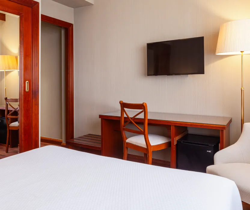 Habitaci&oacute;n de hotel con cama blanca, TV y l&aacute;mpara de pie.