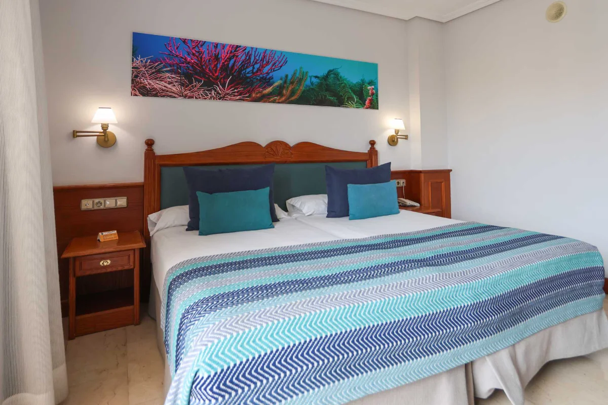 Dormitorio con cama grande, cojines azules y manta a rayas, lámparas y cuadro tropical.