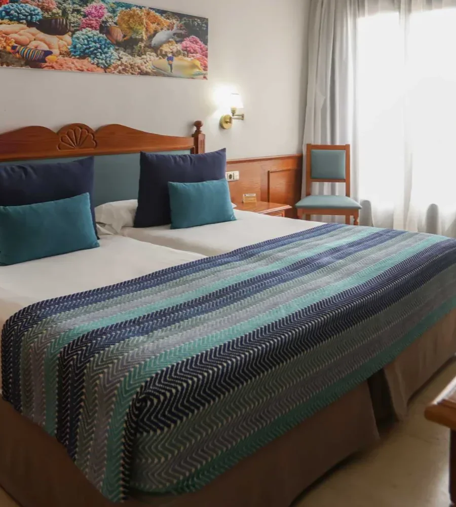 Habitaci&oacute;n hotelera con cama doble y colcha a rayas turquesa y gris