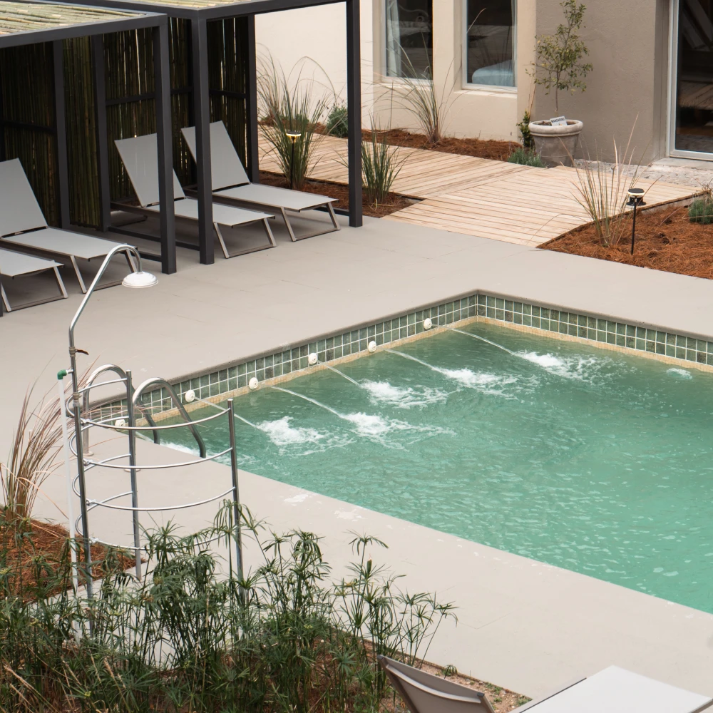 Piscina verde con azulejos, tumbonas, patio y plantas junto a la casa