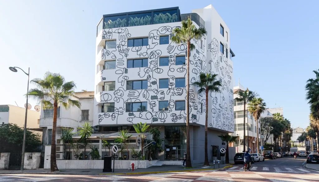 Edificio blanco con mural abstracto y panel de vidrio, palmeras y calle
