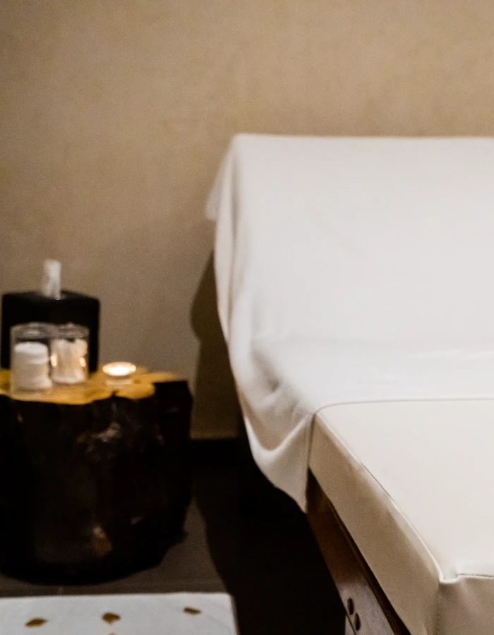 Mesa auxiliar con vela y frasco, junto a una cama con s&aacute;banas blancas