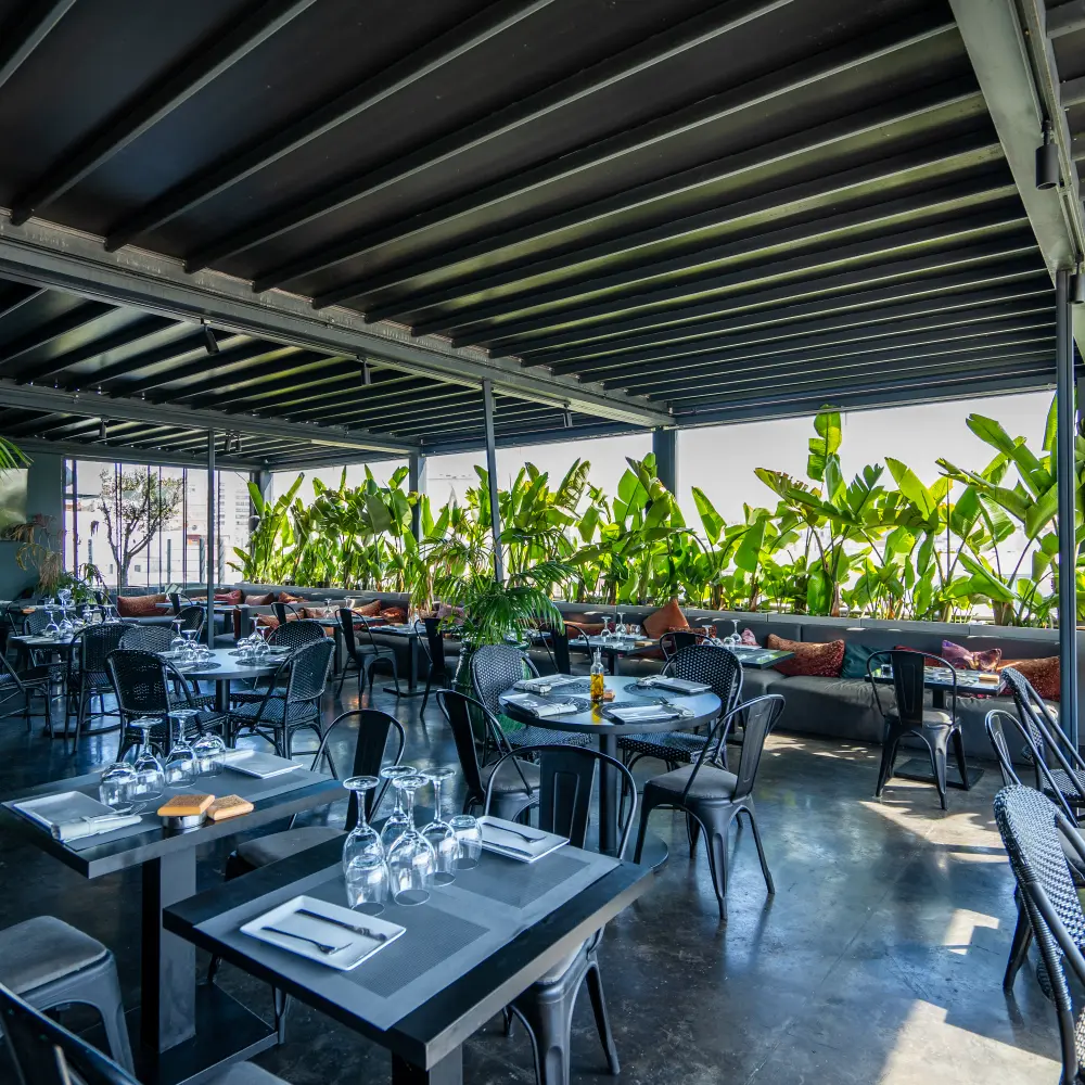 Terraza techada con mesas y sillas, decoraci&oacute;n de plantas verdes.