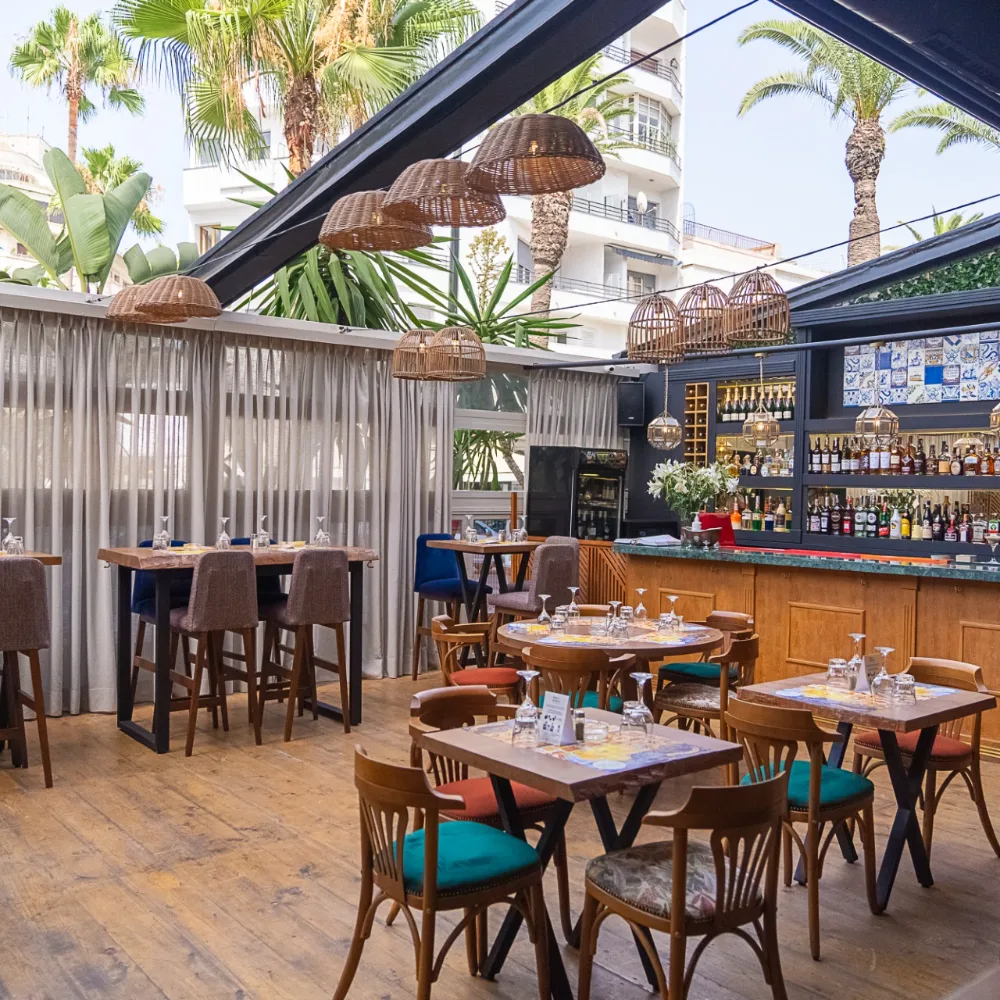 Terraza de restaurante con mesas, sillas, bar y l&aacute;mparas de mimbre bajo palmeras