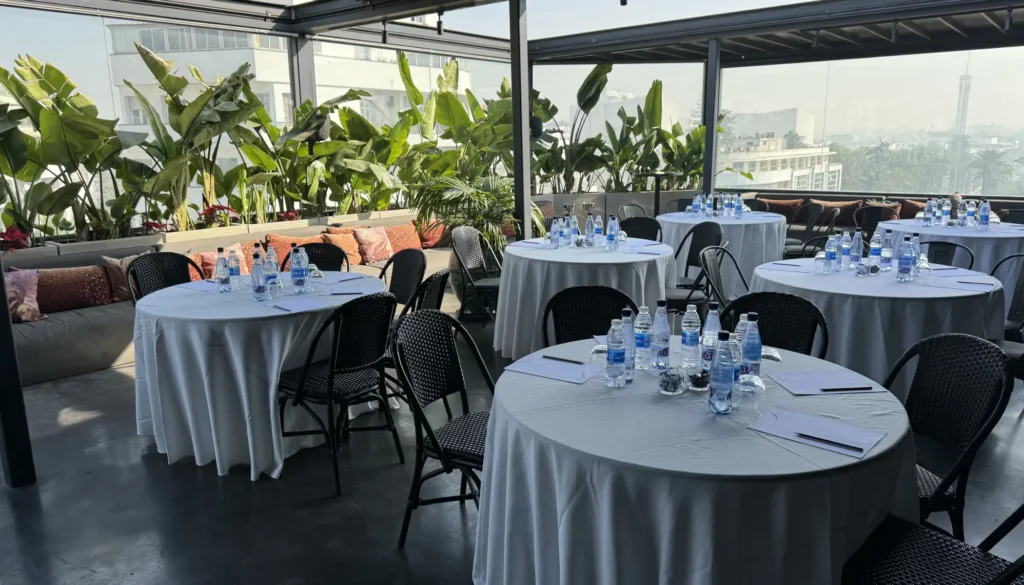 Terraza con mesas para reuni&oacute;n, botellas de agua y plantas, vista urbana.