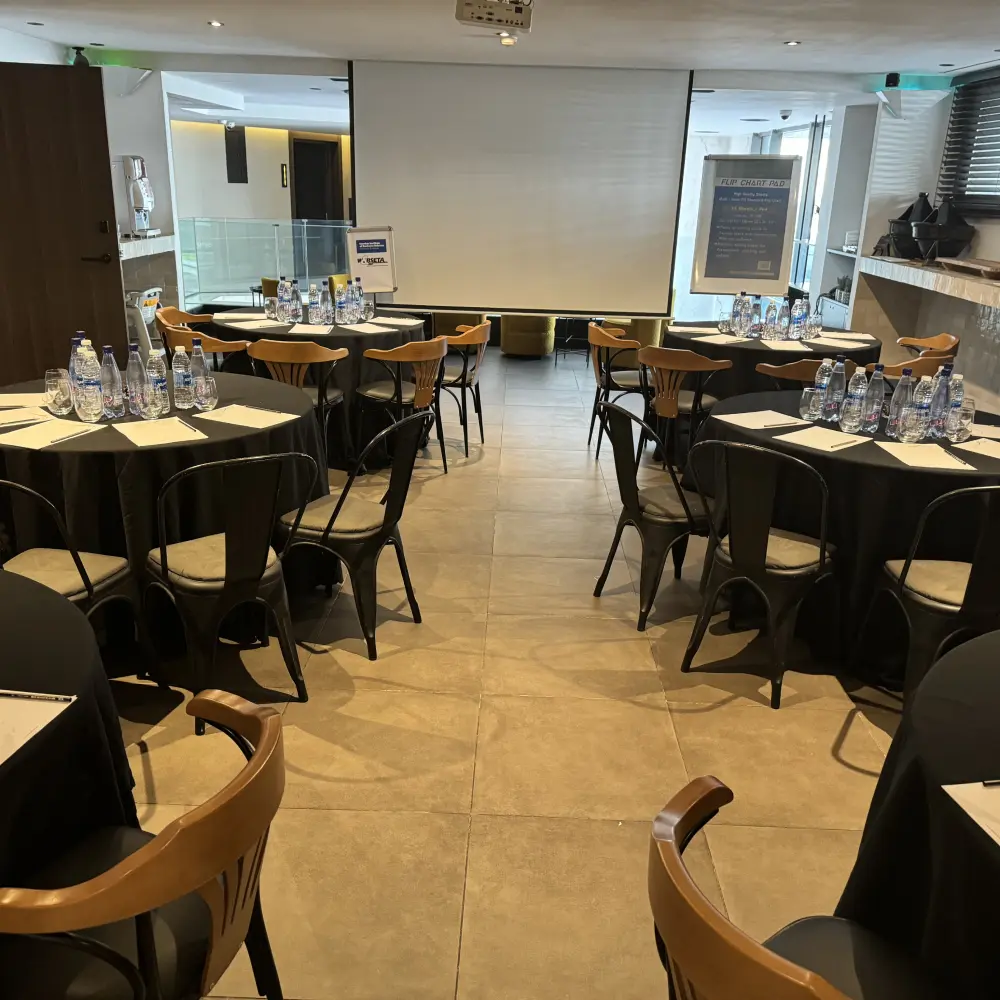 Sala de conferencias con mesas, sillas y botellas de agua, proyector al fondo.
