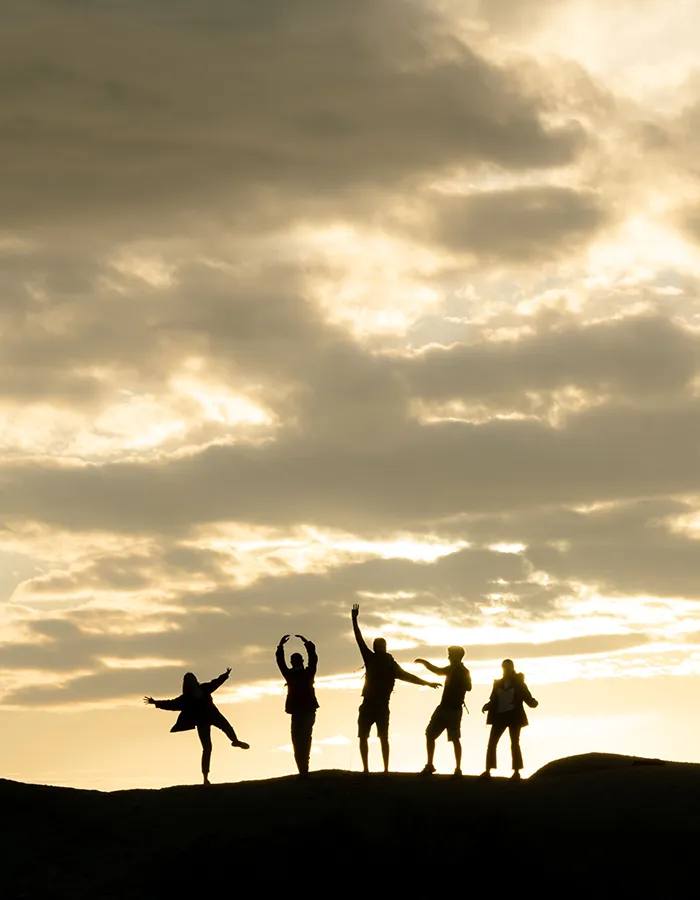 Siluetas de cinco personas bailando en una colina al atardecer, con nubes y sol