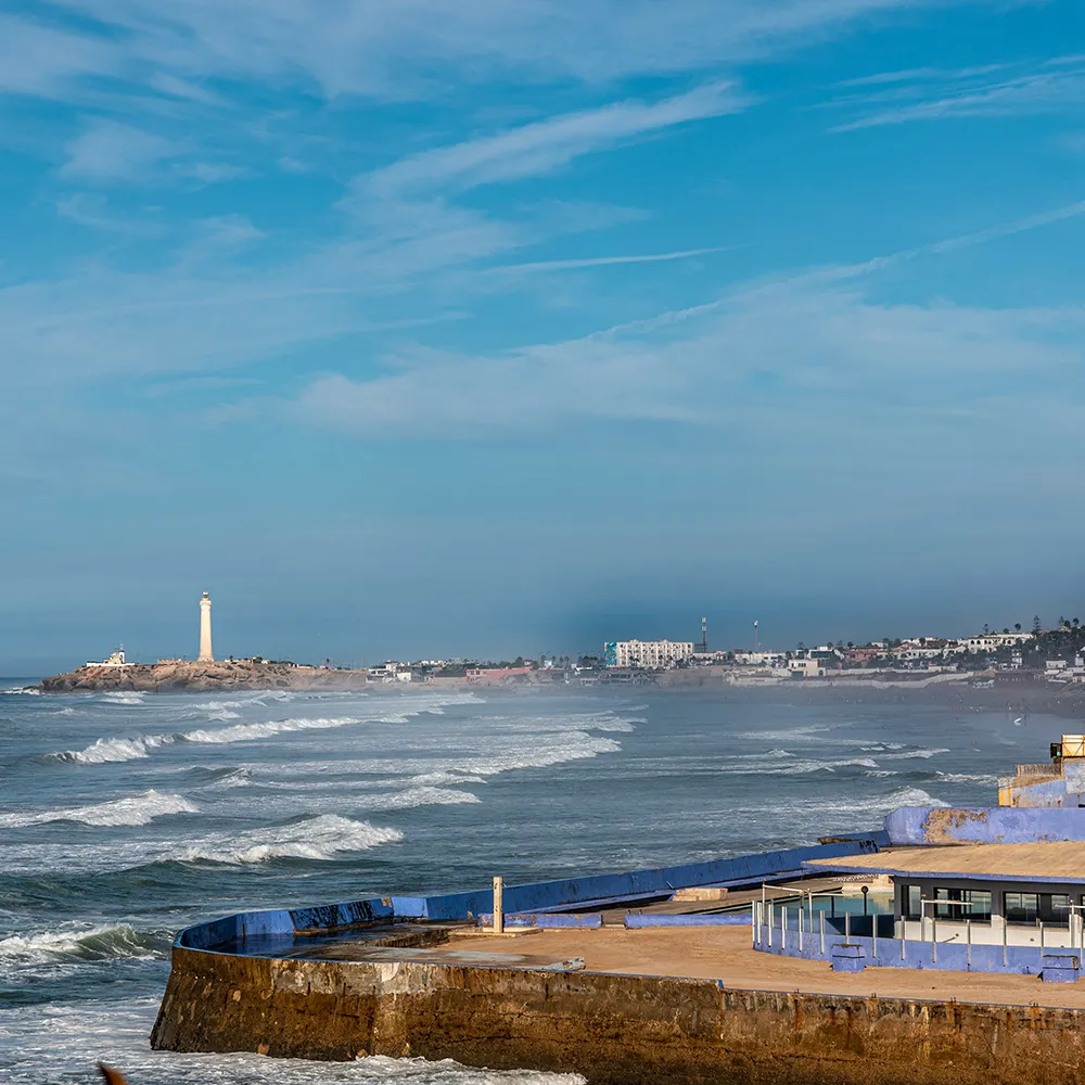 Faro costero y oleaje fuerte contra un muro, cielo azul sobre la playa