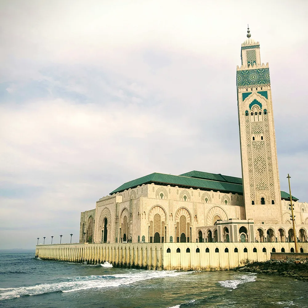 Torre de una mezquita con azulejos verdes y mar al fondo, cielo nublado.