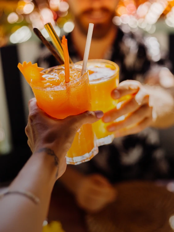 Dos manos brindan con un c&oacute;ctel naranja con hielo y pajitas.
